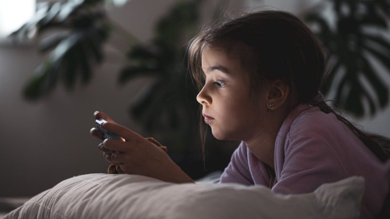 Little girl uses a smartphone lying on the pillow at home.